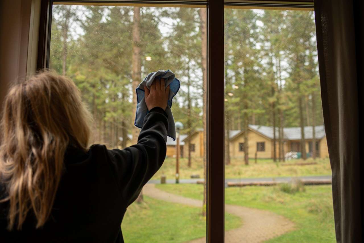 Person wipes a window with a blue cloth; indoors view looking out to a forested area with cabins, curving paths, and grassy grounds under overcast light.