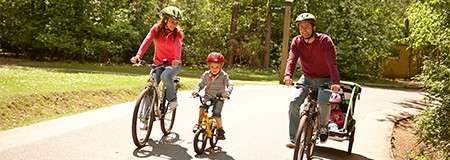 Three cyclists pedal together; a child rides a small bike with training wheels, flanked by two adults, one towing a child trailer; on a sunny, tree-lined park path.