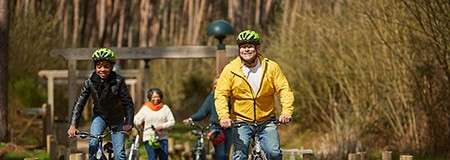 Cyclists pedal forward, smiling, wearing green helmets and casual jackets. They ride along a wooded path with trees and shrubs, wooden posts lining the trail under soft sunlight.