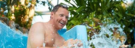 Man laughs while descending a blue water slide, splashing water around him; tropical plants and sunlight surround the slide in a bright aquatic park setting.