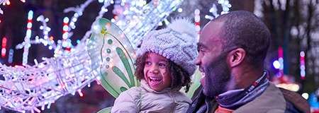 Child wearing fairy wings and a winter hat laughs while being held by an adult, amid colorful festive lights and decorations outdoors at night.