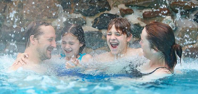 Family of four splashes and laughs, standing chest-deep in a pool as water cascades from above, with bright blue water and a stone wall backdrop, droplets spraying around them.