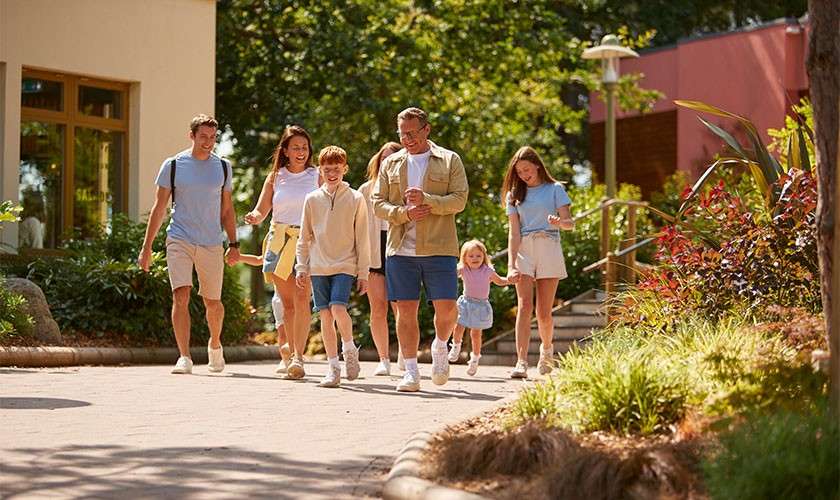 Family group walks and chats, some holding hands, along a sunlit paved path. Surrounding context: landscaped gardens, trees, and low buildings; a window displays the word SALON.