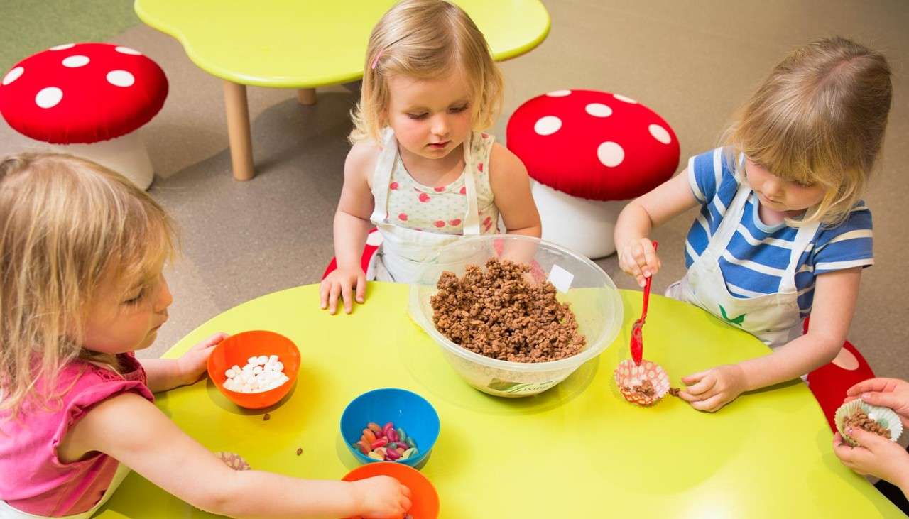 Three toddlers sat round a table with bowls of chocolate crispies and toppings.