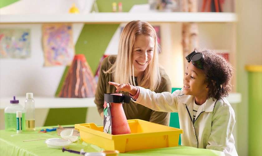 Child in lab coat reaches toward a model volcano in a yellow tray; adult supervisor smiles nearby; classroom setting with shelves, paintings, and science supplies in the background.