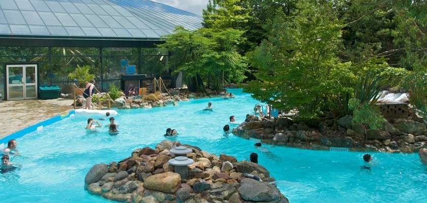 Swimmers float and wade through a turquoise, winding pool, navigating around rock islands and lush trees; a glass-roofed building and poolside steps sit beside them in a landscaped leisure complex.