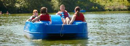 Blue pedal boat carries four people wearing life jackets, pedaling and relaxing on calm lake water, while green, sunlit trees line the shore in the background.