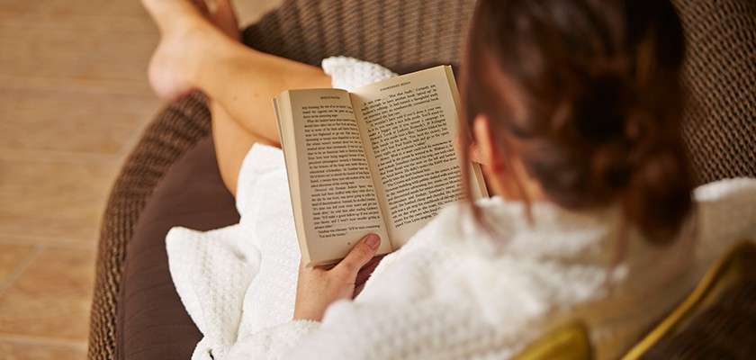 Book—held open and read by a person in a white robe—while reclining on a wicker lounge chair, legs crossed, in a warm, tiled indoor setting.