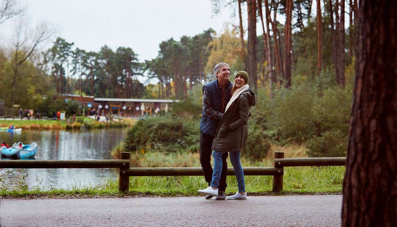 Couple walks arm-in-arm, smiling, on a paved path beside a lake. Forest of tall pines and a lodge with people and kayaks in the background; wooden fence lines the path.