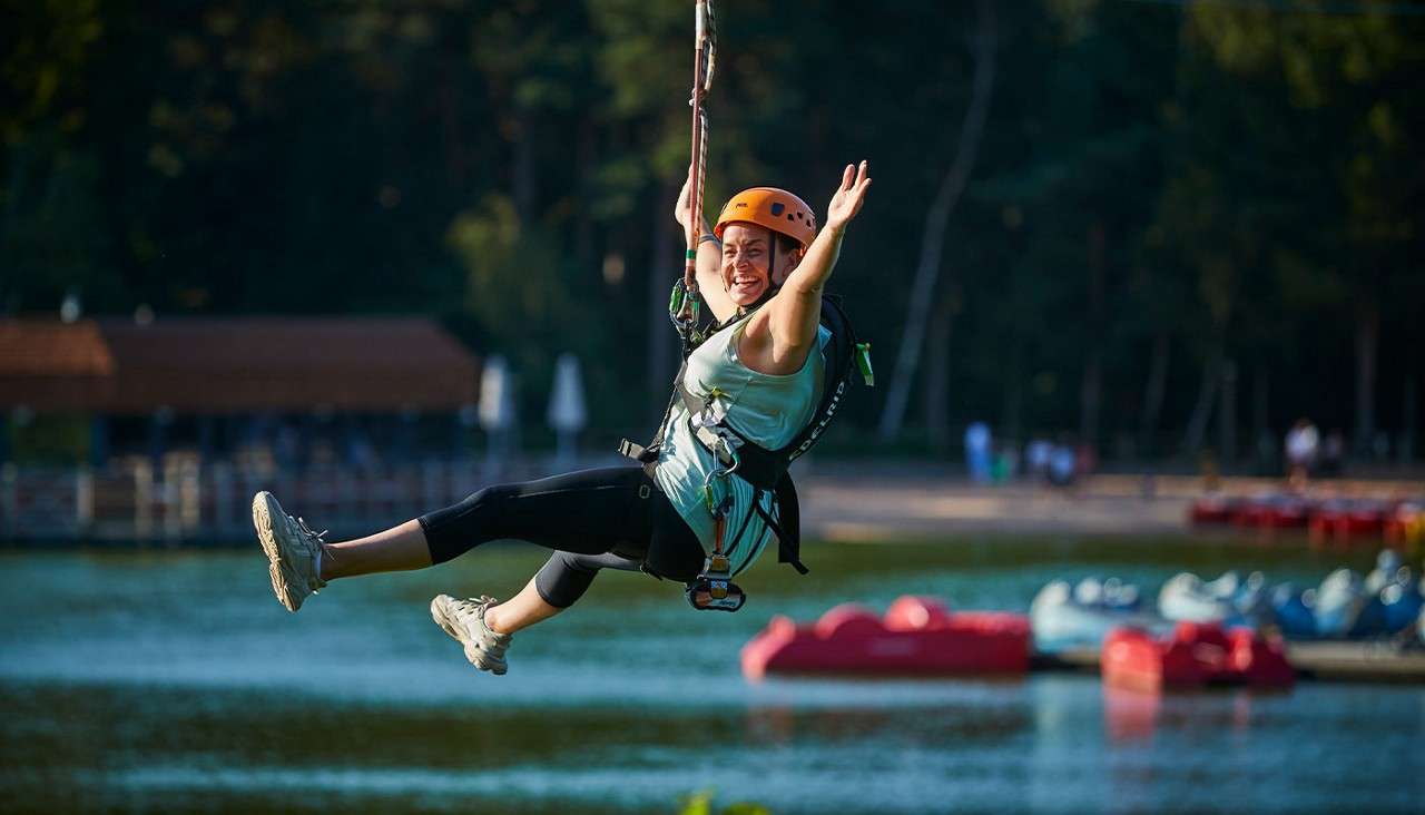 A harnessed zip-line rider glides smiling with arms outstretched, legs forward; above a lakeside park with dock, paddle boats, and a forested shoreline in soft evening light.