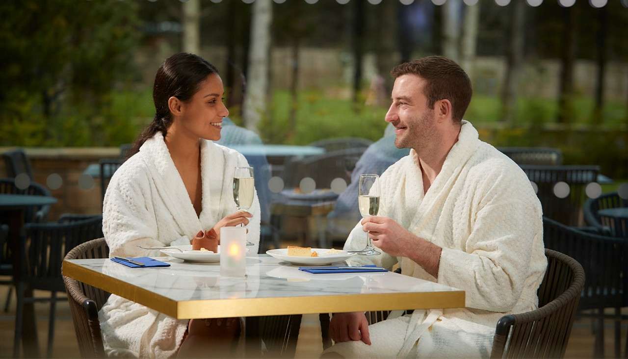 Two adults in white bathrobes chat while raising champagne flutes, seated at a marble-topped table with plates and a candle, on an outdoor café patio bordered by trees and glass.