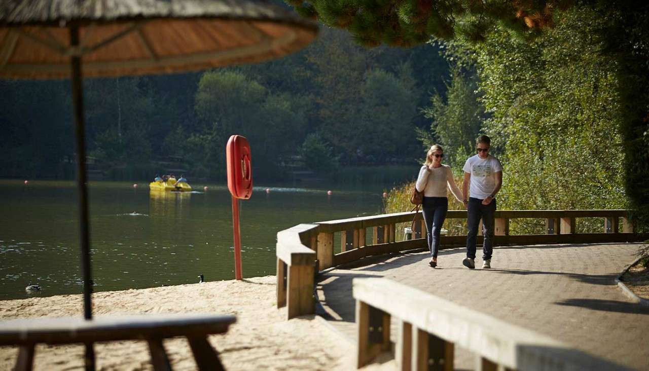 Couple walks hand-in-hand along a lakeside path, bordered by wooden railings. Nearby, a sandy beach, lifebuoy post, and ducks frame calm water where people pedal a yellow boat amid forested banks.
