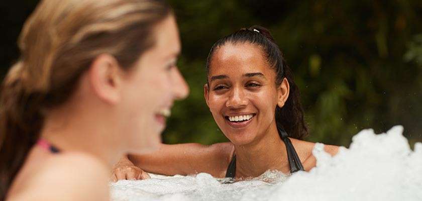 Two people relax and chat, smiling in a bubbling hot tub; one leans on the edge, foam swirling, with green foliage blurred in the background, indicating an outdoor setting.