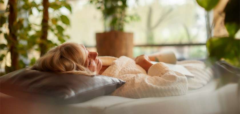 Person reclines on a cushioned lounge, eyes closed, wearing a textured white robe and resting on a dark pillow, surrounded by indoor plants and soft daylight in a tranquil spa-like room.