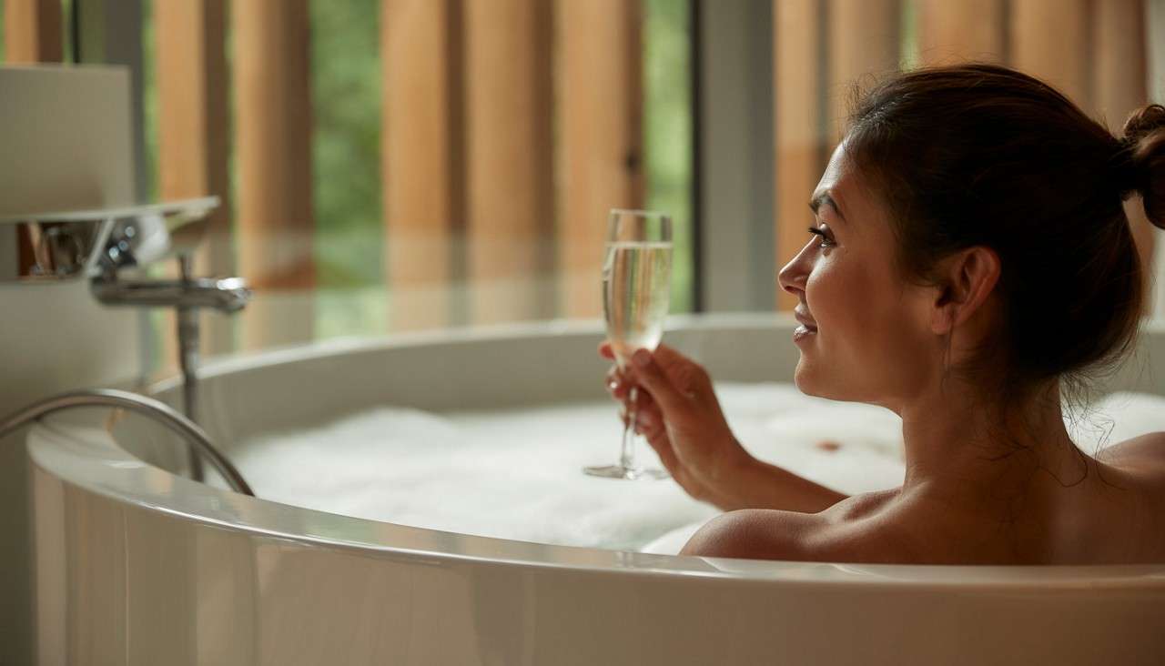 Person holds a champagne flute, reclining in a round bubble bath. Context: modern bathroom with chrome faucet beside them and tall vertical wooden window slats letting in soft daylight.
