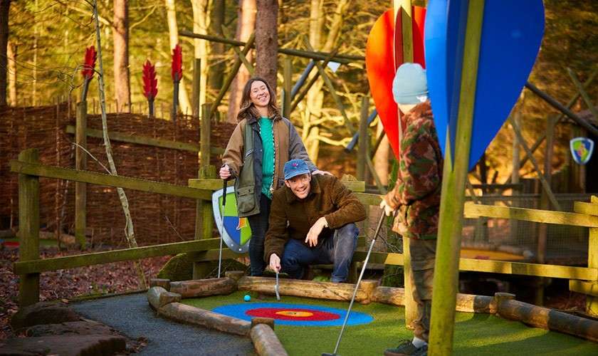 Man putts a blue ball on a colorful target while two companions watch and hold clubs, within a wooded mini-golf course bordered by wooden rails and large decorative leaves.