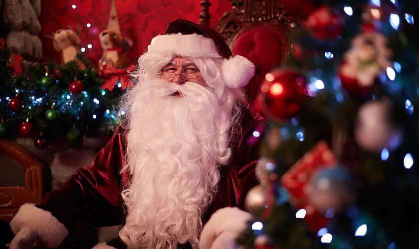 Person dressed as Santa Claus sits smiling on a red armchair, white-bearded and velvet-suited, surrounded by a lit Christmas tree, ornaments, and garlands in a warmly decorated festive room.
