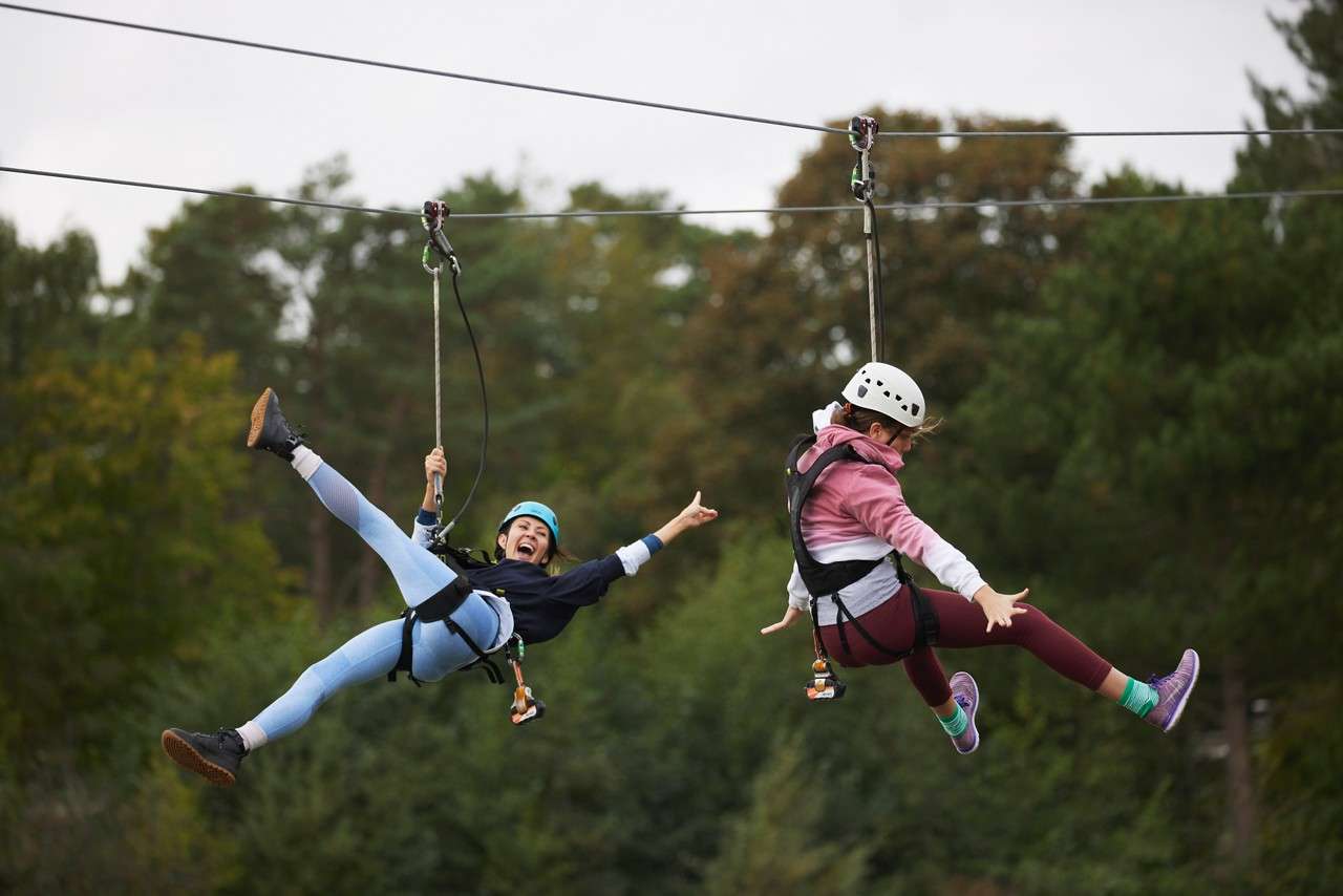 Two helmeted riders glide on parallel ziplines, legs outstretched and arms spread, suspended in harnesses, against a backdrop of tall green trees and overcast sky in an outdoor adventure park.