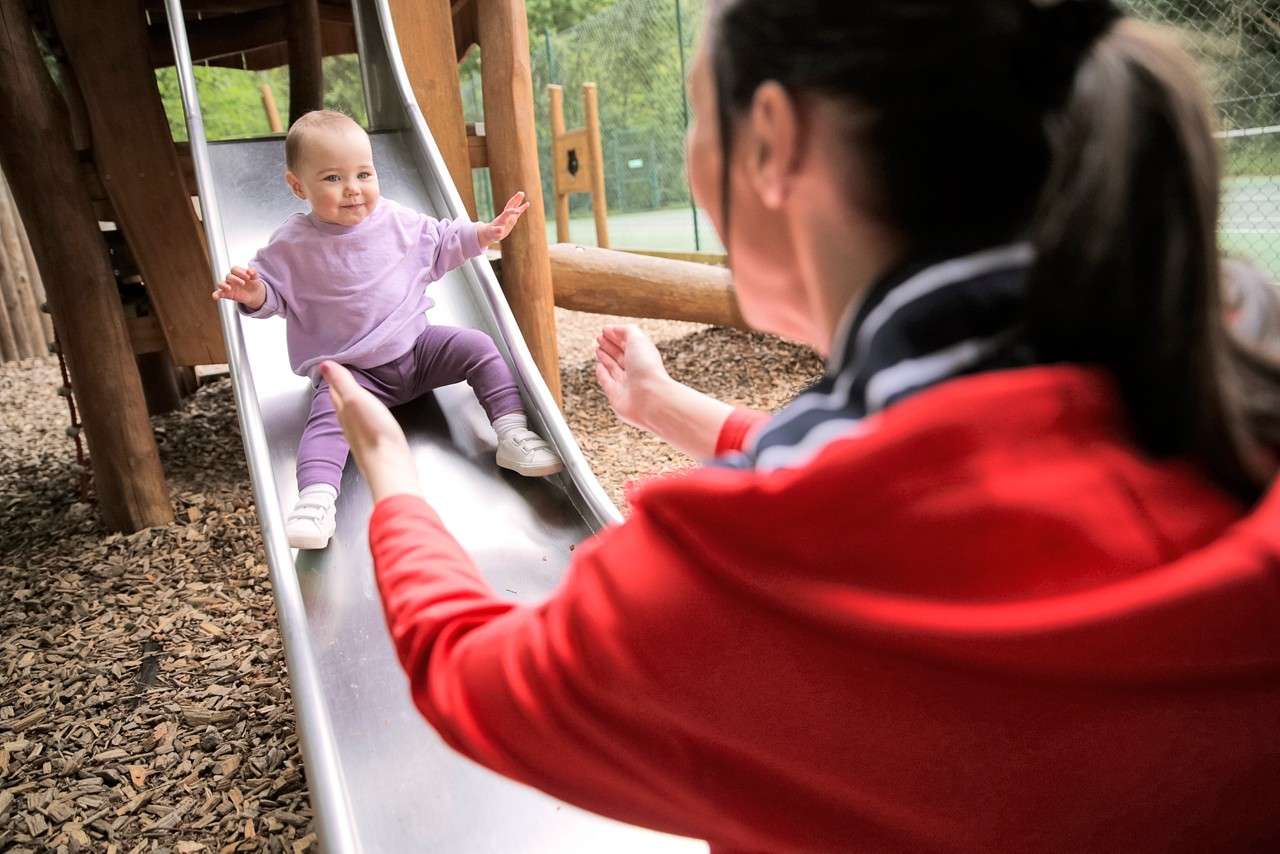 A baby slides down a metal playground slide, arms out, while an adult in red reaches to catch them amid wood-chip ground, wooden play structure, and fenced greenery.