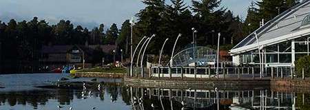 Glass-walled lakeside pavilion reflects on calm water, bordered by curved metal arches and a promenade railing; seagulls float nearby while pine trees and wooden lodges line the opposite shore.