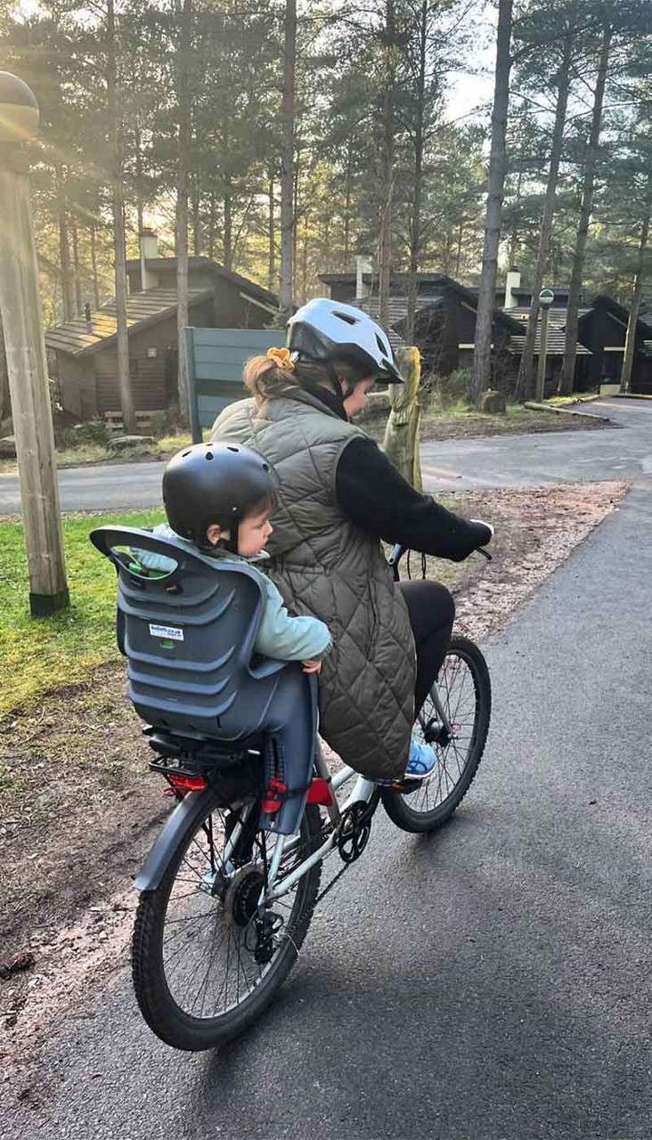 Adult cyclist carries a toddler in a rear child seat, both wearing helmets. They ride along a paved path through a wooded cabin area with tall pines and soft sunlight.