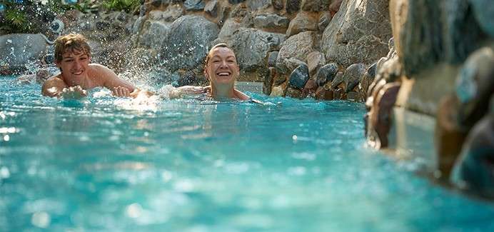 Two swimmers smile and paddle, splashing water, as they move through a turquoise pool bordered by natural stone walls and greenery, suggesting a resort-style or landscaped water feature.