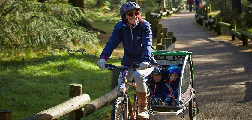 Adult cyclist pulls two children in a covered bike trailer, riding along a shaded forest path with wooden railings; all wear helmets and jackets as sunlight filters through the trees.