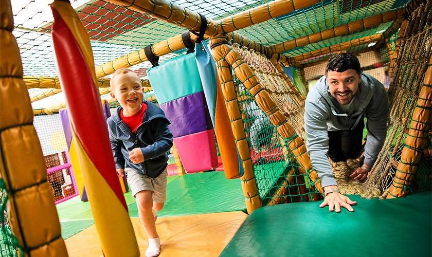 Child runs ahead while an adult crawls through a net tunnel, both smiling; context: colorful indoor soft-play structure with padded beams, hanging foam blocks, and safety netting.