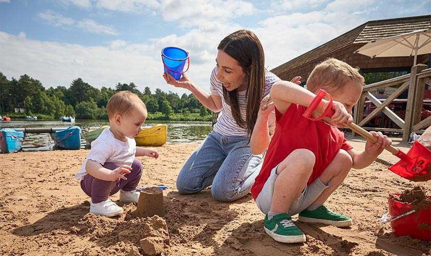 Children build sandcastles as a woman assists, using buckets and red shovels; they crouch on a sandy lakeside beach near kayaks and cabins under a partly cloudy sky.