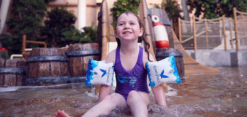 Child splashes while sitting in shallow water, smiling; wearing purple swimsuit and inflatable armbands; small slide, barrels, buoy, and rope fence in an indoor water play area.
Text: Confidence (on both armbands)
