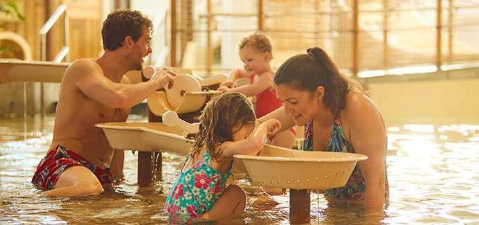 Parents and two young children play with water tables, pouring and scooping in a shallow indoor pool. Warm lighting, wooden railings, swimsuit attire, gentle splashing, smiling in an interactive play area.
