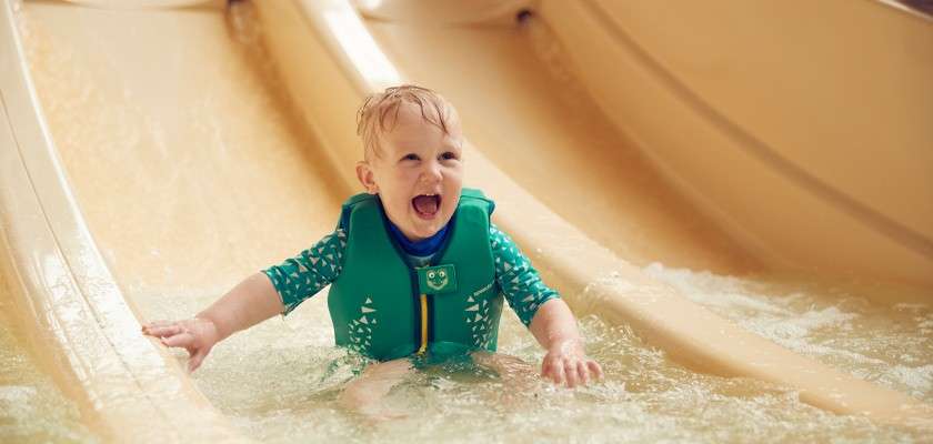 Toddler splashes and laughs, sitting upright after descending a slide, wearing a green life jacket. Context: shallow water at the base of a beige water slide in an indoor pool.