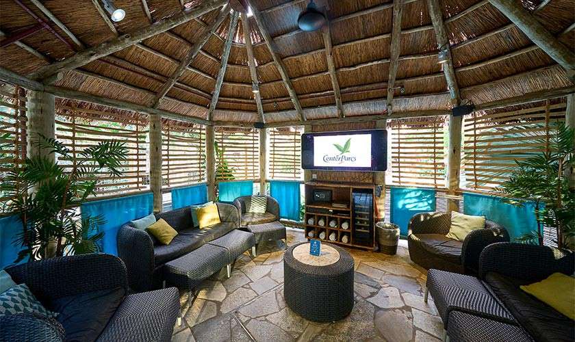 Curved wicker sofas encircle a round ottoman, facing a wall-mounted TV displaying “Center Parcs,” inside a thatched-roof gazebo with wooden slat walls, stone floor, and potted tropical plants.
