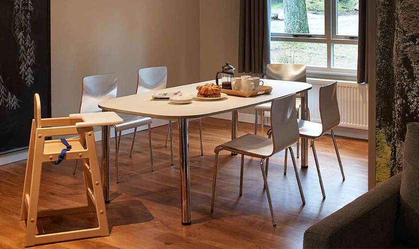 Dining table holds a tray with coffee press, cups, and croissants; surrounded by four metal chairs and a wooden high chair in a bright, wood-floored room with window and curtains.