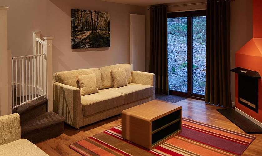 Beige sofa faces a low wooden coffee table, resting on a striped rug. Surrounding context: curtained glass doors to a rocky yard, orange corner fireplace, staircase with baby gate, and forest-photo wall art.