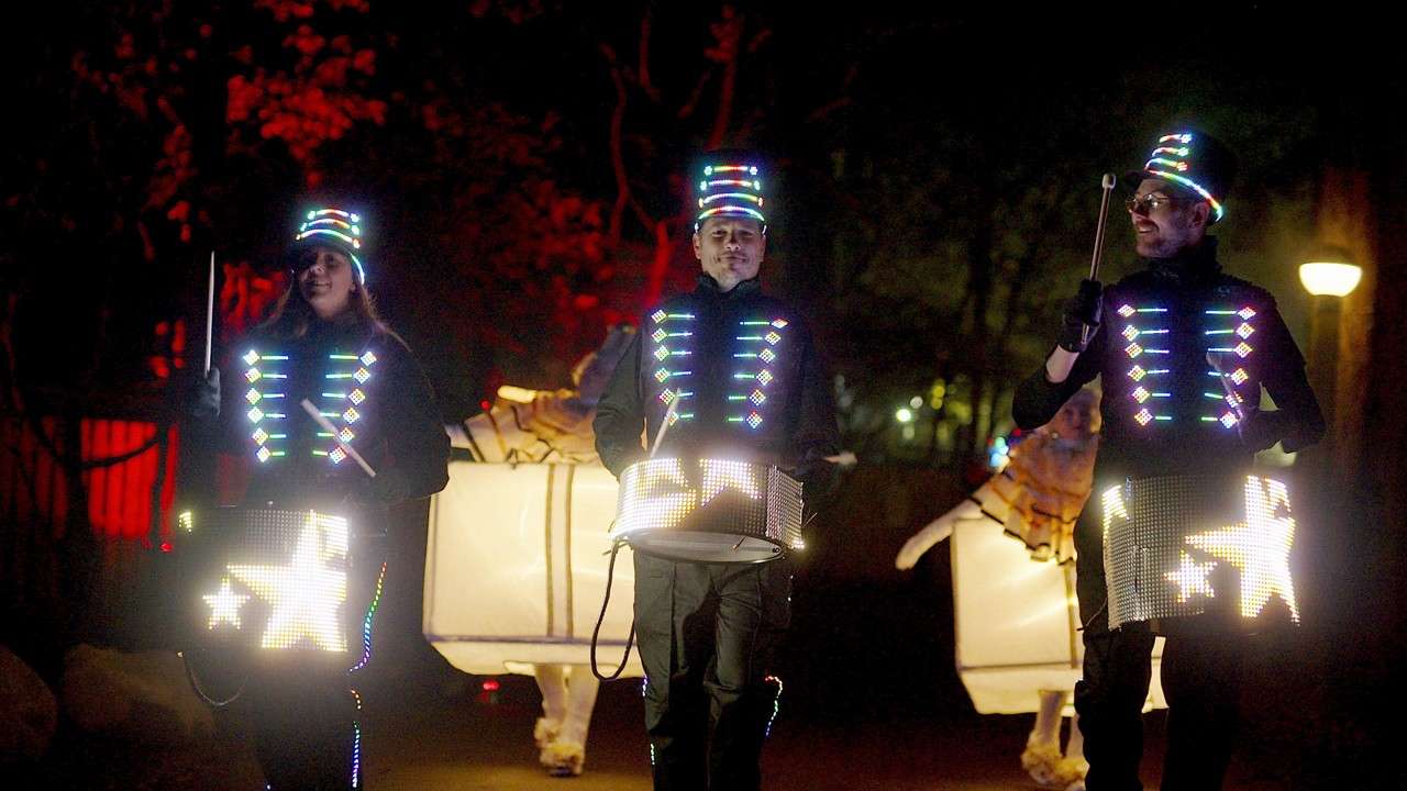 LED-clad drummers march and play illuminated drums, their vests and hats glowing. Behind them, performers in lighted animal costumes follow along a dark pathway with red-lit trees and lampposts.