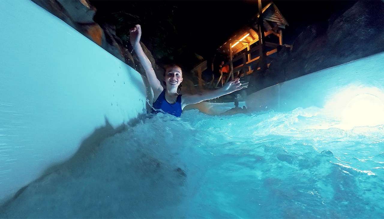 Person rides a water slide, arms raised, smiling, as churning blue water splashes around; white slide walls curve under a dark setting with a warmly lit wooden platform overhead.