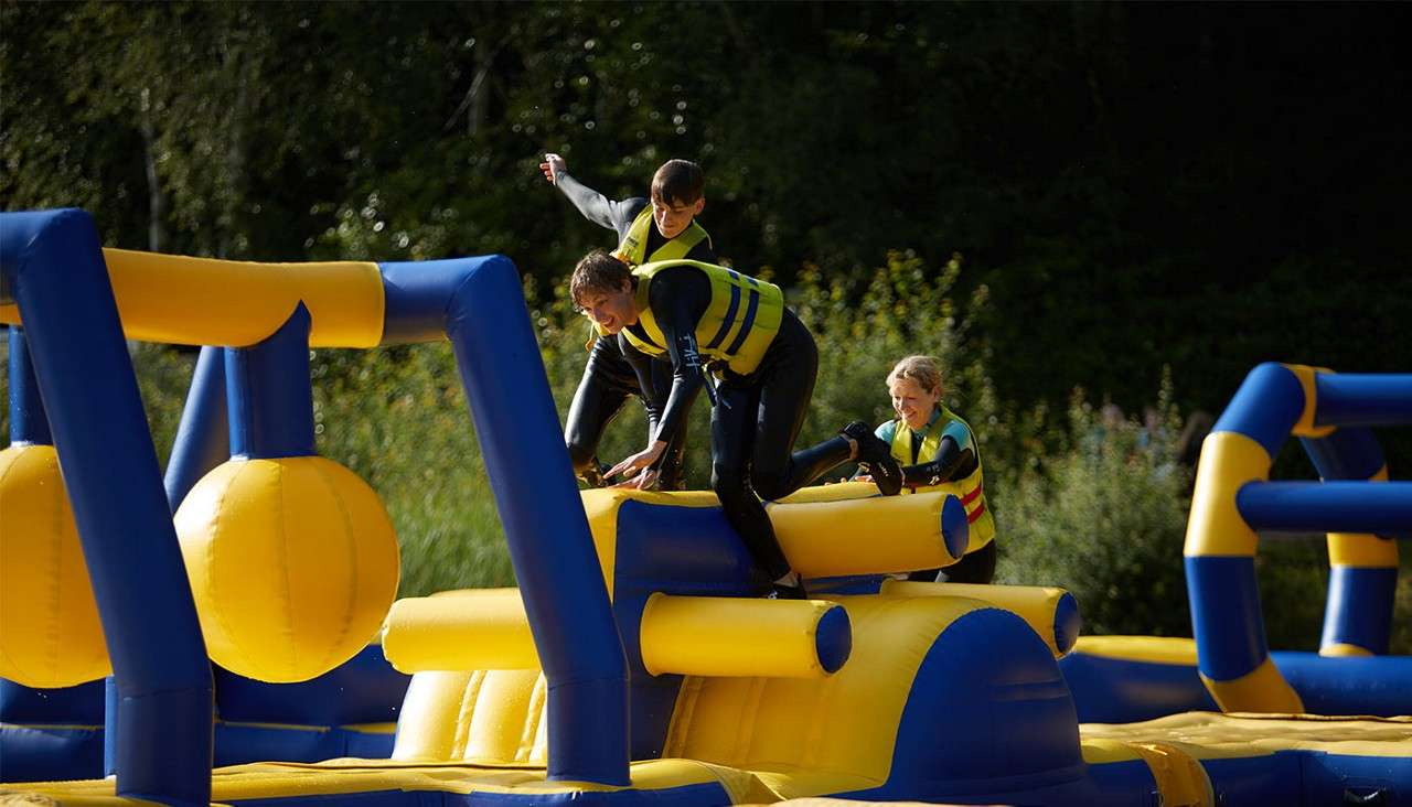 Three life-jacketed participants climb and balance across a yellow-and-blue inflatable obstacle, dodging hanging balls, in an outdoor waterpark set beside greenery under daylight.