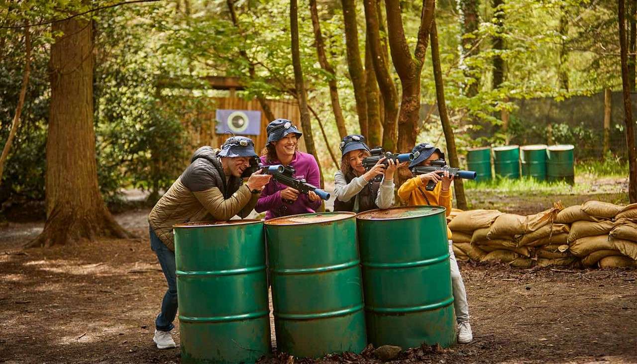 Four players aim laser-tag blasters while crouching behind three green barrels; in a wooded arena with sandbags, trees, and a target board in the background.