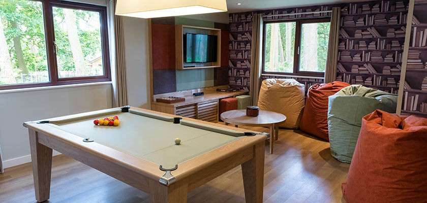 Pool table holds a racked set of balls, ready for play; surrounding lounge includes beanbag chairs, round coffee table, wall-mounted TV, windows with forest view, and bookshelf-pattern wallpaper.