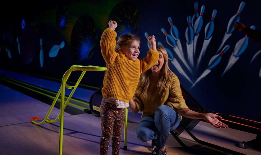 A little girl on a bowling lane cheering with her arms in the air.