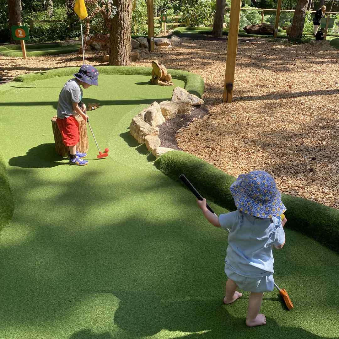 Two children with putters play on a mini-golf hole. One lines up a red ball; the other approaches. Trees, rocks, a frog statue, and a sign reading “2” nearby.