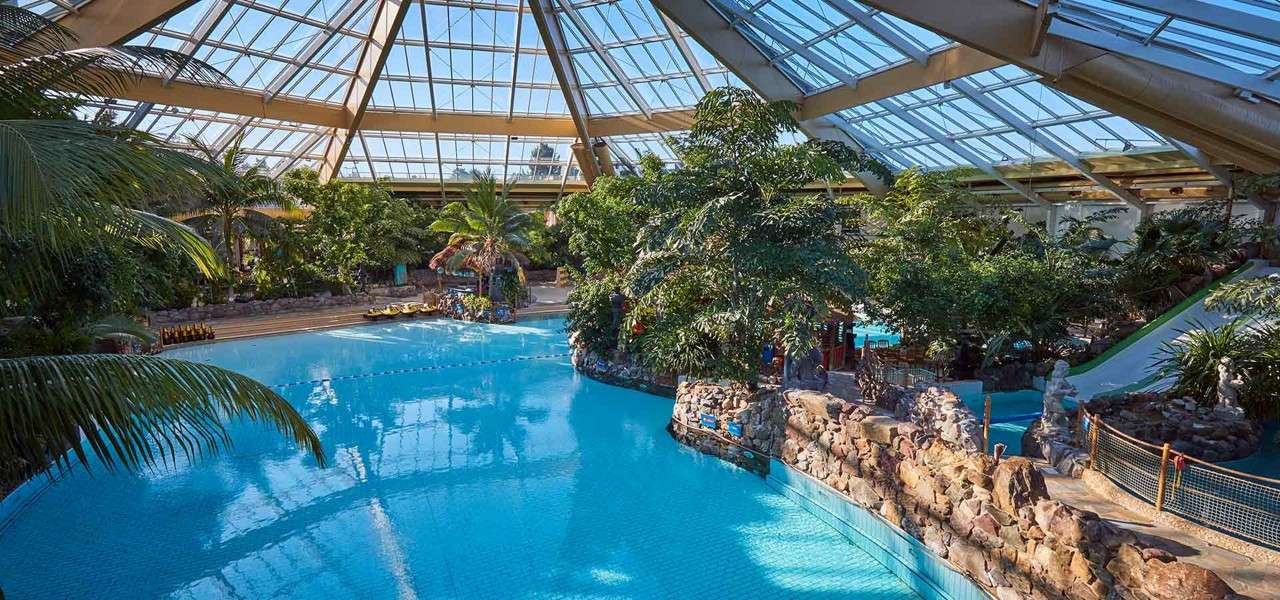 Swimming pool stretches and curves around rocky islands, reflecting skylights; surrounded by tropical plants, palm trees, lounge chairs, and a waterslide under a large glass-domed roof.