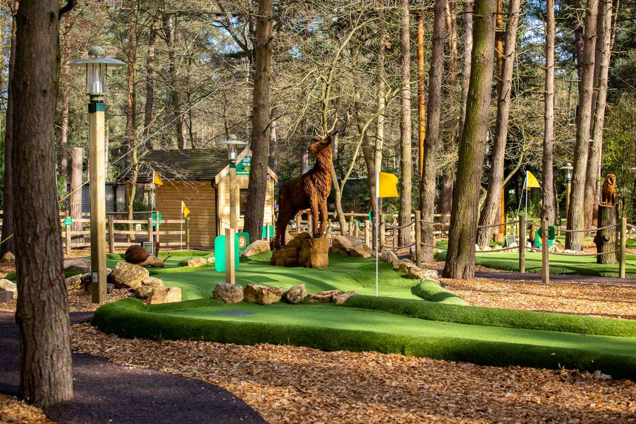 Mini-golf course displays yellow-flagged holes; a carved stag stands on rocks. Surrounded by tall trees and a wooden hut. Visible text: EXIT, 9.