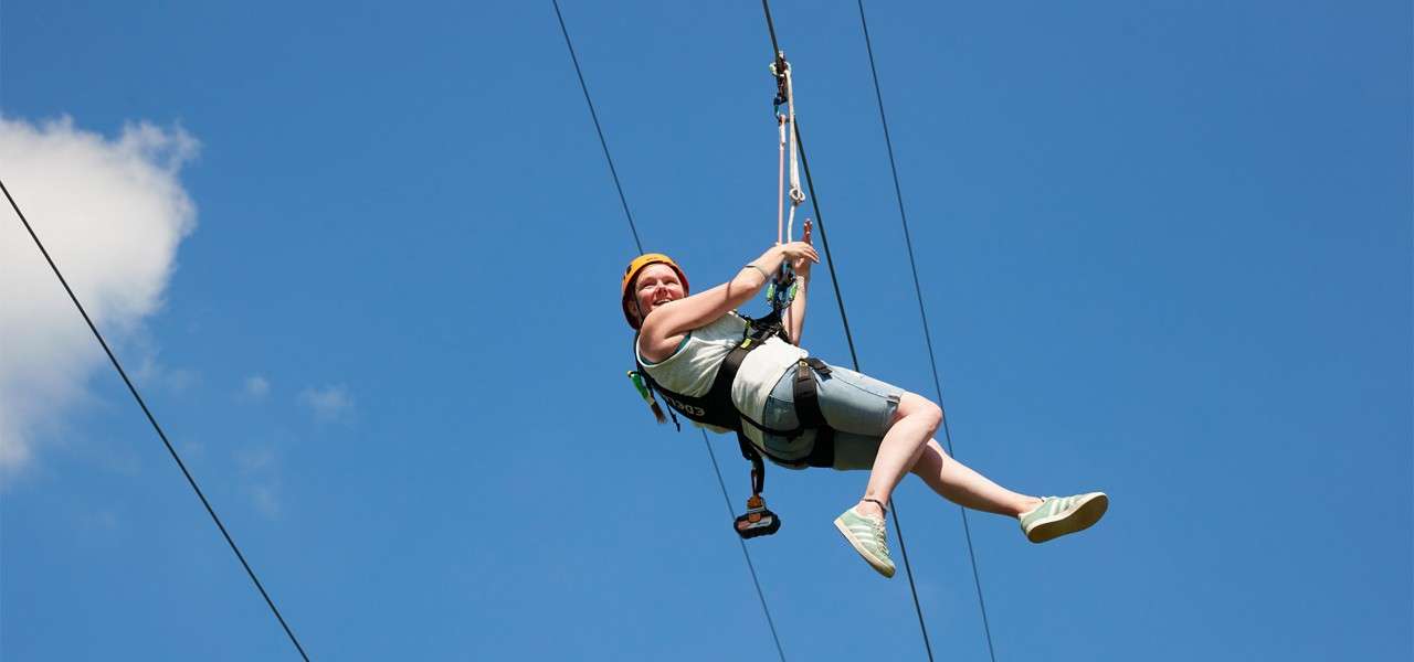 A person wearing a helmet and harness glides on a zip line, legs extended, gripping the trolley, against a clear blue sky with cables stretching diagonally.