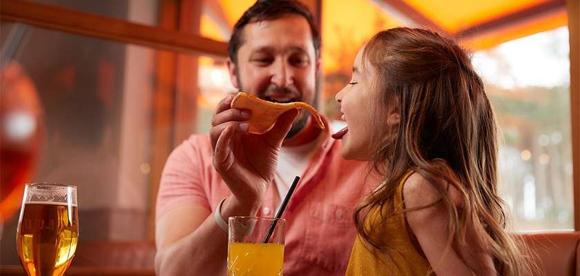 Man holds a pizza slice, feeding a child; she leans forward with mouth open. They sit at a restaurant table with an orange drink, a beer, and warm orange lighting.