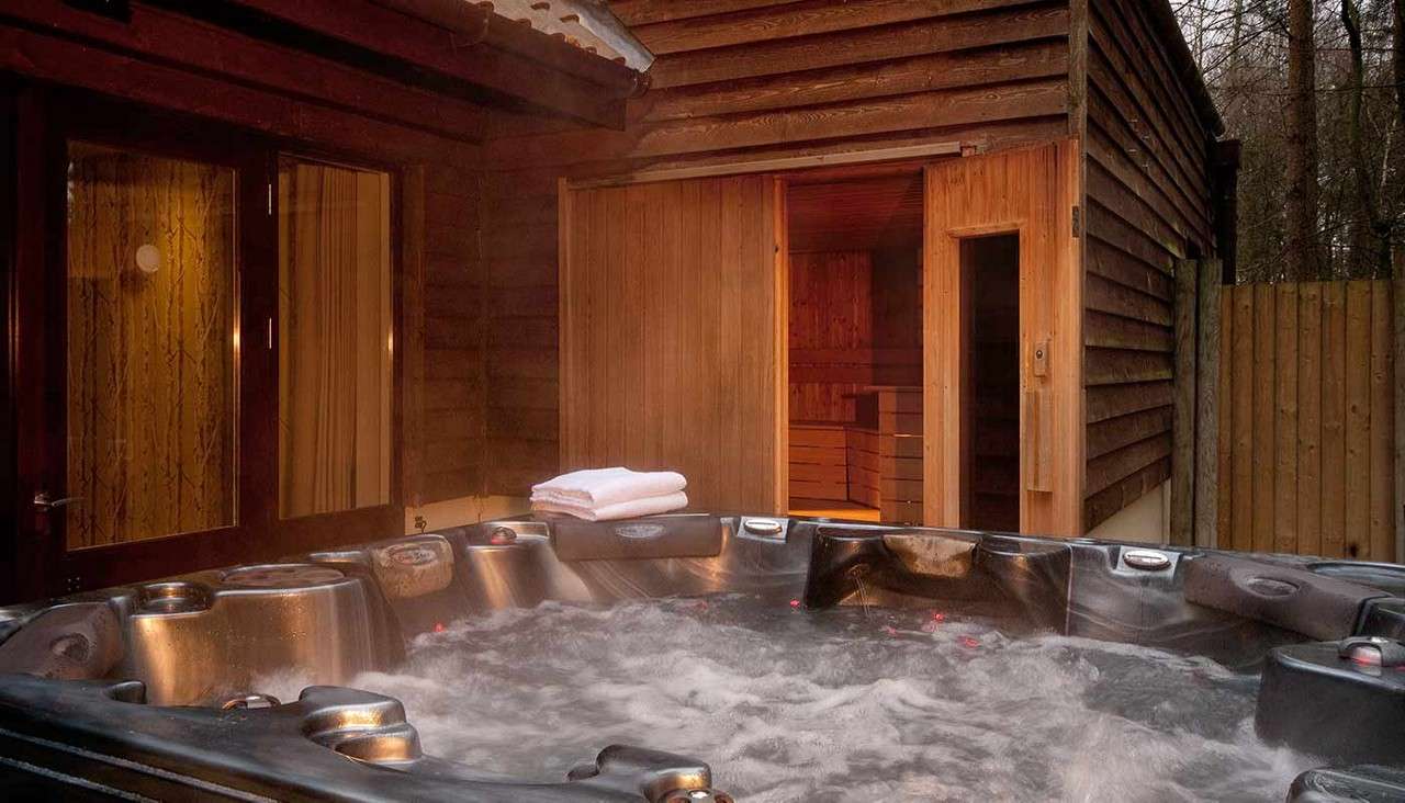 Hot tub bubbles vigorously, steam rising; folded white towels rest on its edge. A wooden cabin with glass doors and an open, warmly lit sauna stands outside among trees.