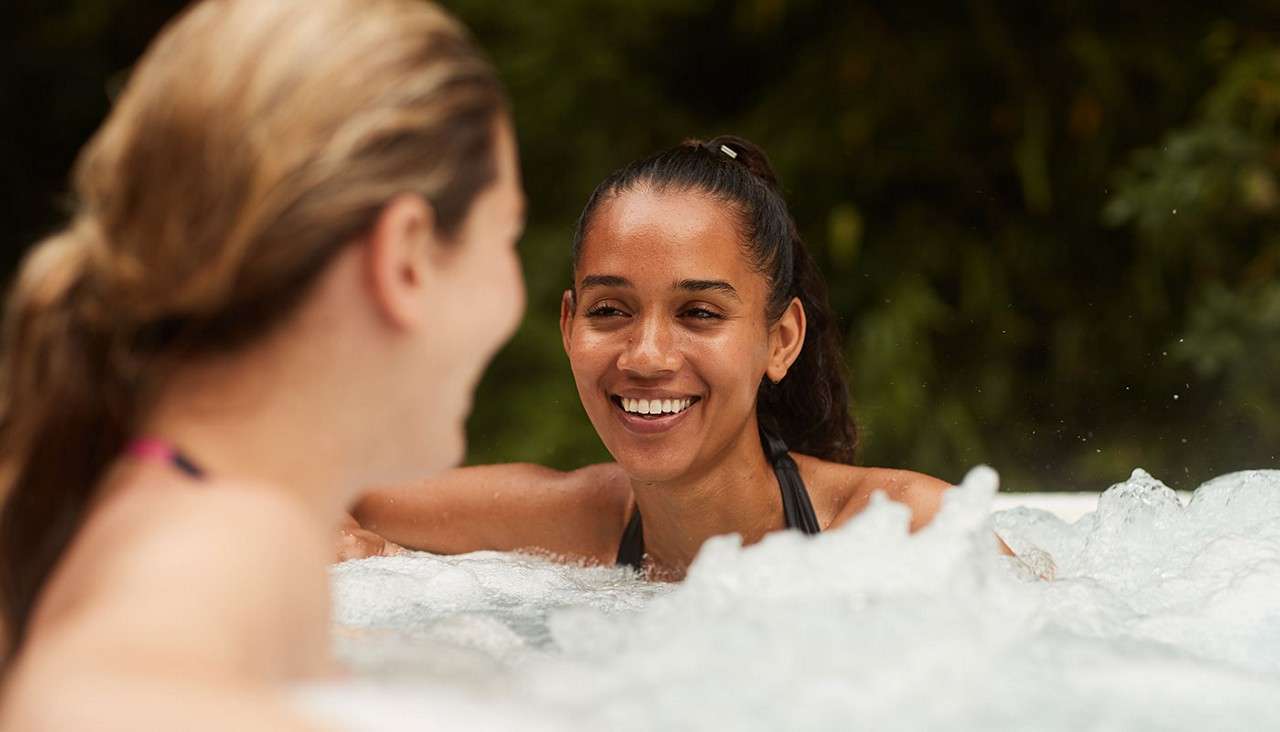 Smiling woman chats while relaxing in a bubbling hot tub, arm resting on the edge; another person faces her in the foreground. Surrounding greenery suggests an outdoor setting.