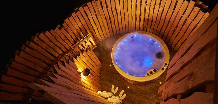 Hot tub bubbles with blue LED lights, steaming. Sitting on a wooden deck, encircled by curved slatted fencing, lit by warm lamps, with robes and slippers placed nearby at night.