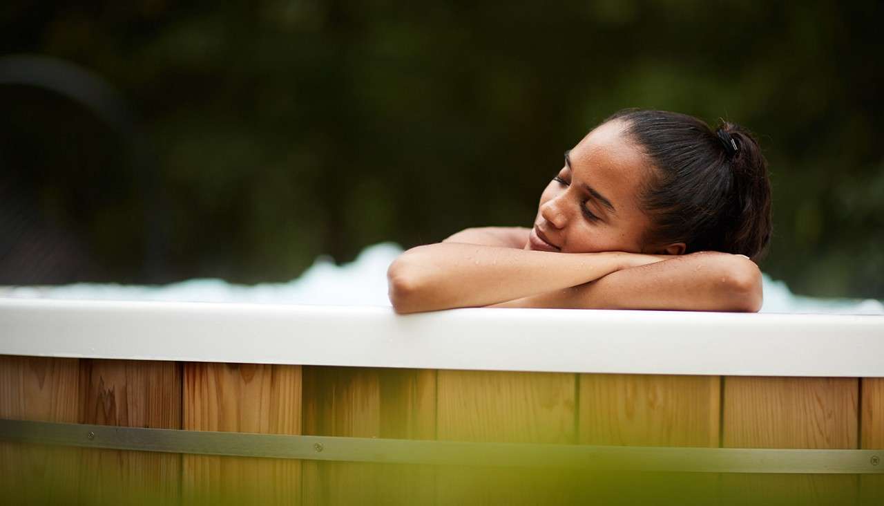 Person in a round wooden hot tub rests arms on the white rim, eyes closed, relaxing; bubbles surround them as blurred green foliage forms the outdoor background.